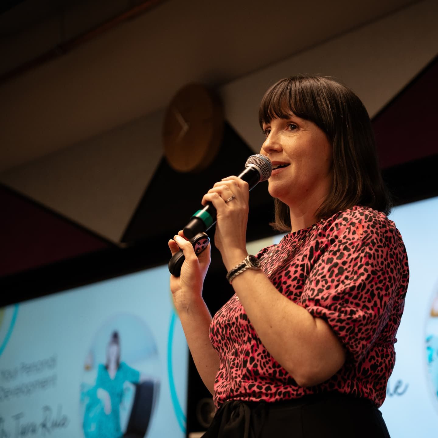 Tara Rule speaking on stage, holding a microphone and presenting during a leadership development event.