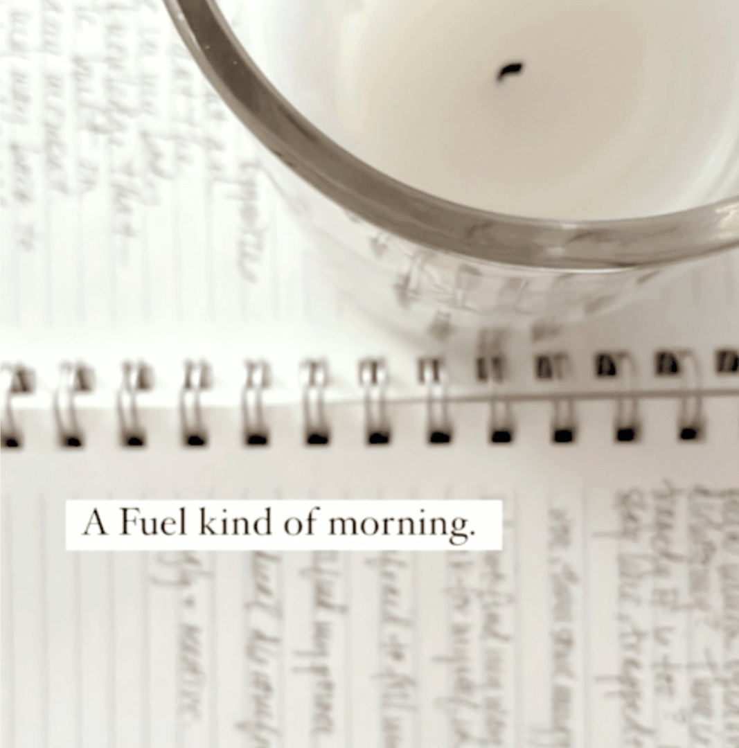 A top-down, macro photograph of an open spiral notebook filled with elegant handwriting. A clear glass candle holder with a lit wick rests on the page, casting a warm shadow over the journal. A clean white text box in the center reads: A Fuel kind of morning.