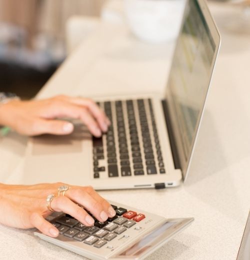 A close-up of a person's hands using a laptop and a calculator simultaneously on a light-colored desk, highlighting a focused workspace for financial planning or business management.