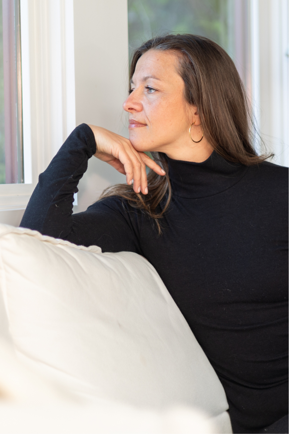 Steffi Seefeld, transformational somatic coach, seated on a white sofa, wearing black, and looking out the window
