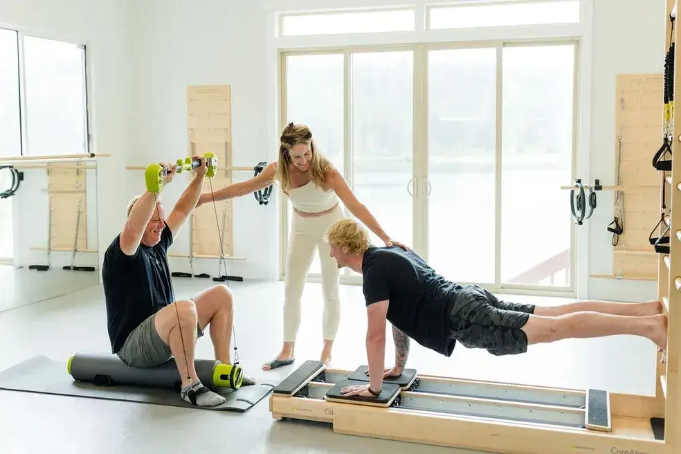 A woman in a white leggings and a white top guides two men through pilates exersizes