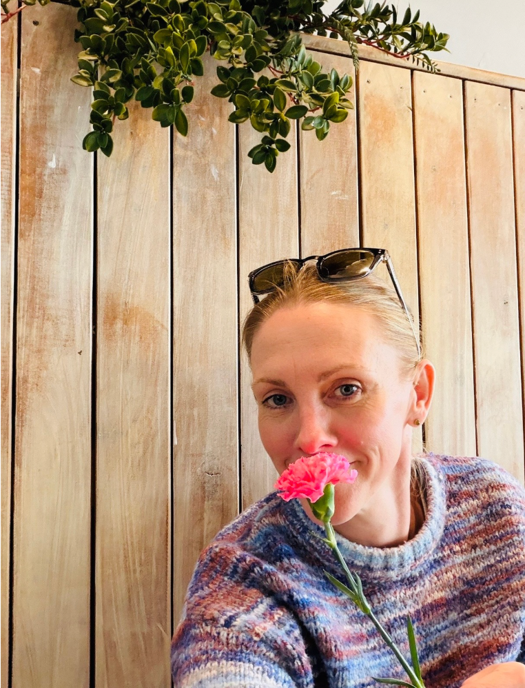 Alison Balson wears a pink sweater, standing with her arms crossed leaning against a white wall