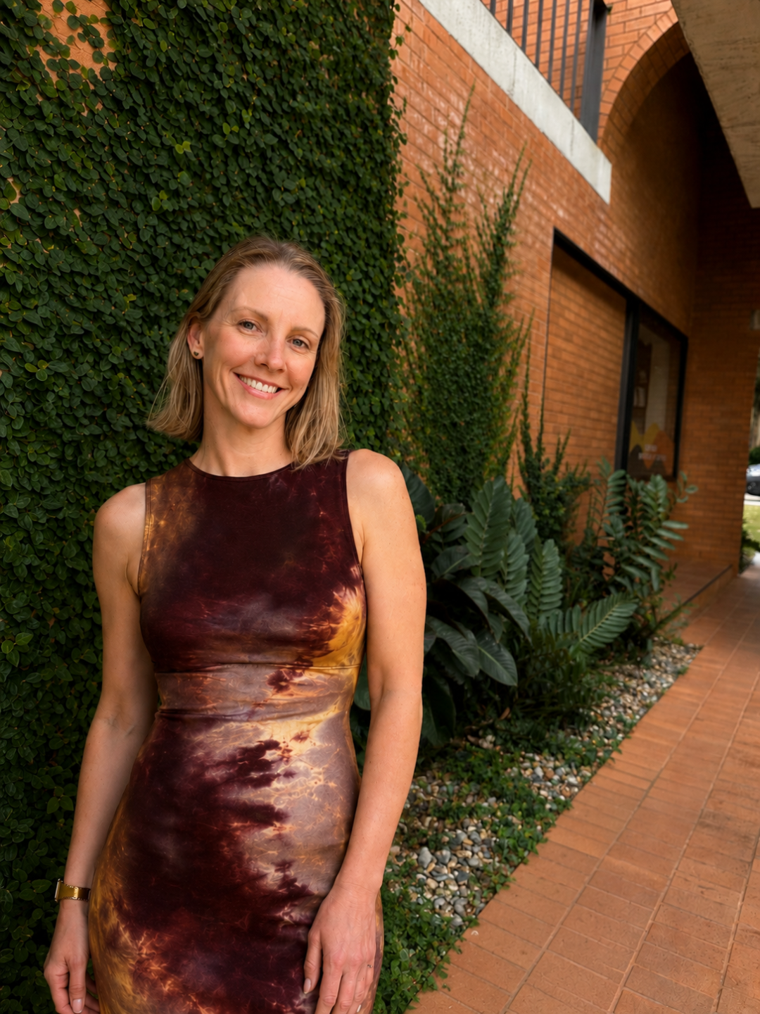 Alison Balson sits in front of a white wall with her arms resting on her knees, wearing a green floral dress smiling at the camera