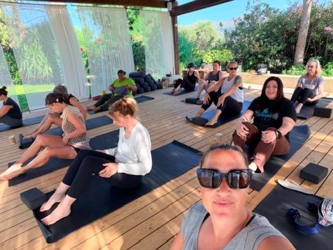 A group of women participating in an outdoor pilates or yoga session on a wooden deck surrounded by lush greenery, with natural light filtering through sheer curtains.