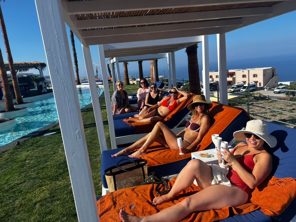 A group of women relaxing on sun loungers by a poolside cabana, with palm trees and a stunning blue Mediterranean sea view in the background.
