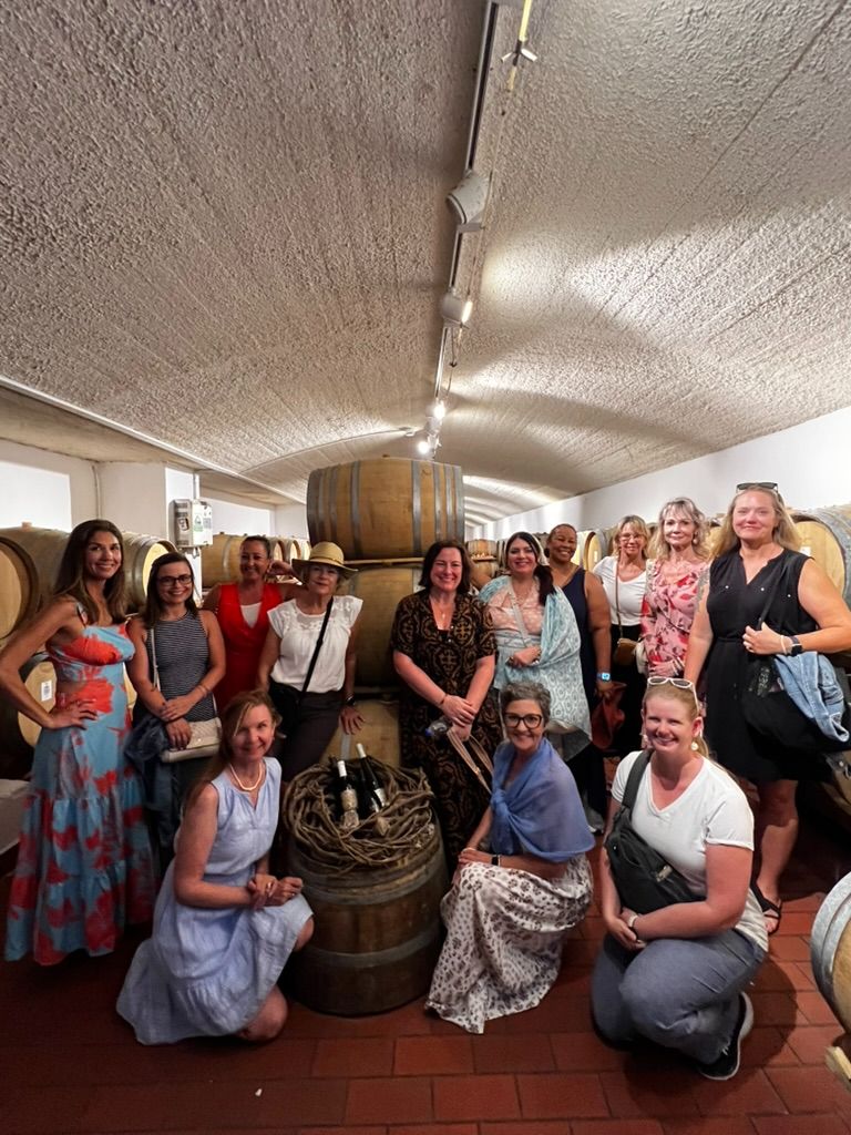 A group of women posing together inside a rustic wine cellar filled with oak barrels, smiling and enjoying a winery tour experience in Mallorca.
