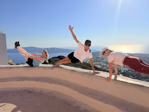 Three women performing pilates exercises on a scenic hilltop overlooking a stunning Mediterranean coastline and blue sea under a clear sunny sky.