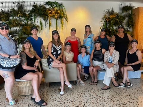 A group of women posing together inside a stylish hotel lobby adorned with tropical greenery, all wearing matching branded caps, dressed in summer outfits.