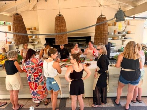 A group of women gathered around an open-air kitchen counter with a chef, surrounded by fresh ingredients, rustic woven pendant lights, and lush greenery during a cooking class experience.