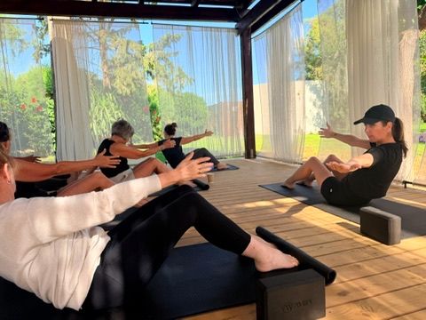 A group of women practicing pilates on reformer beds inside an elegant open-air pavilion surrounded by lush greenery and natural light.