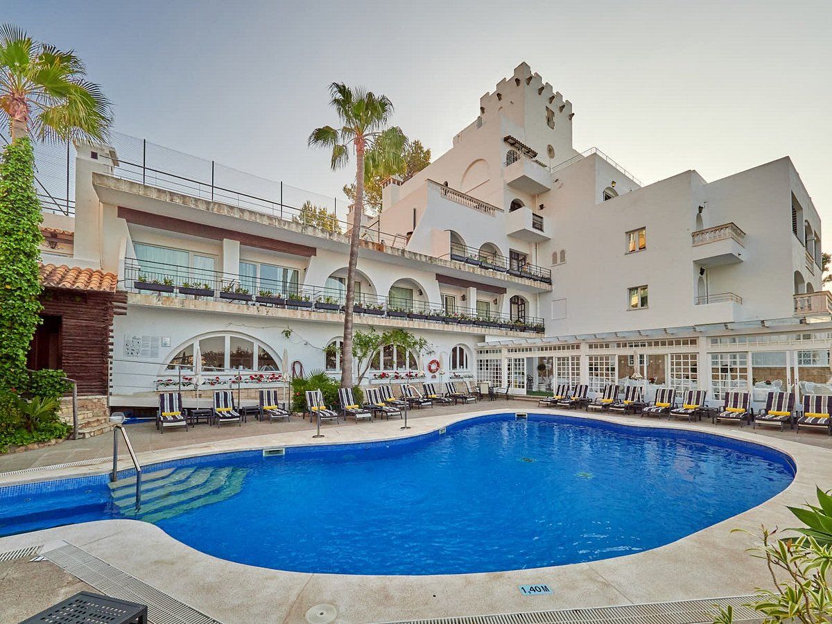 Exterior view of a elegant white Mediterranean-style hotel featuring a stunning oval swimming pool, sun loungers, arched balconies, and palm trees at dusk in Mallorca.