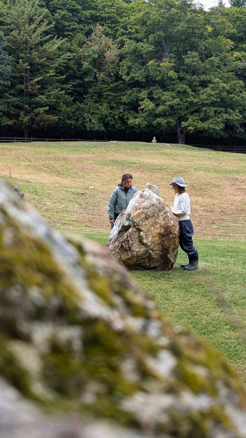 two women standing around a stone in an open field