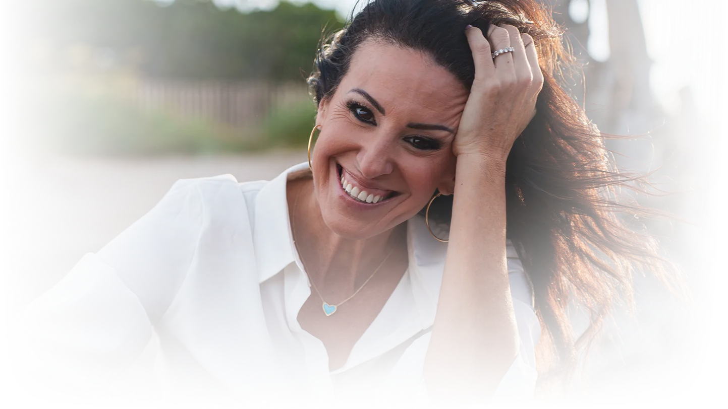 A woman with long brown hair smiles warmly, holding her hair back with her hand. She wears a white blouse, hoop earrings, and a heart-shaped necklace.