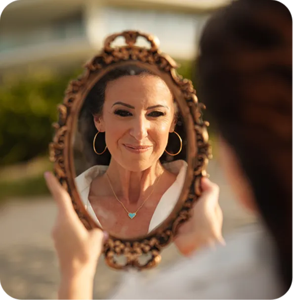 A woman smiles at her reflection in an ornate oval mirror. She wears gold hoop earrings and a heart-shaped necklace. The background is blurred greenery.