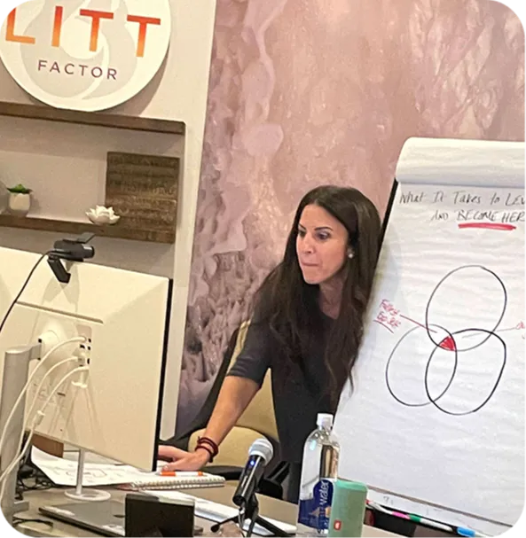A woman is leading a discussion, seated at a desk with a computer and water bottle. A flip chart shows a Venn diagram. The setting is an office.
