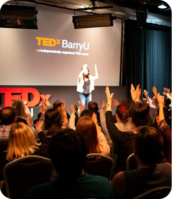 Audience with raised hands faces a speaker on stage at TEDxBarryU. The speaker is mid-presentation, evoking an engaging and interactive atmosphere.