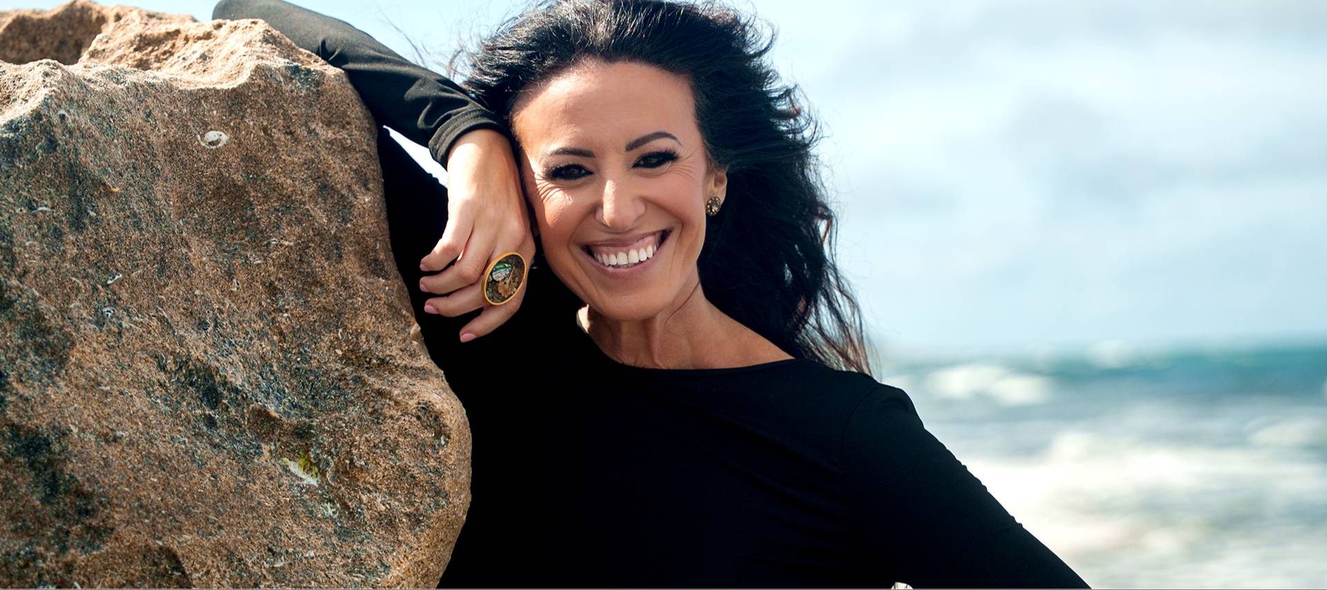 Smiling woman in black top leans against a large rock by the sea. Her hair is windblown, and she wears a large ring. The mood is joyful and serene.