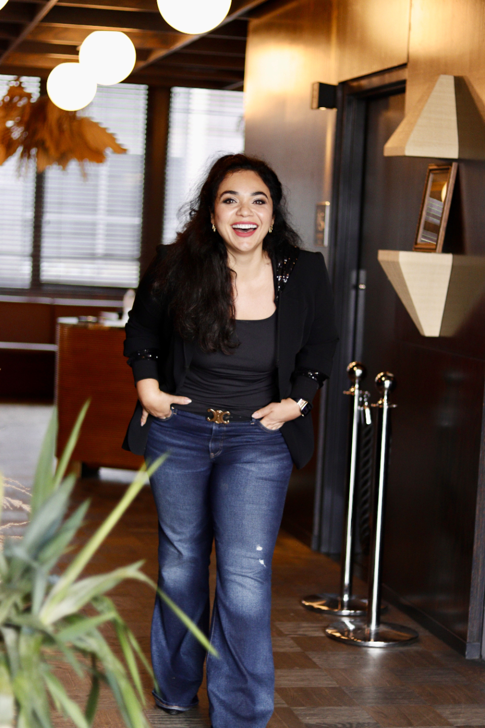 Dunya Daikhi laughing in a café, jeans and black blazer, red lip