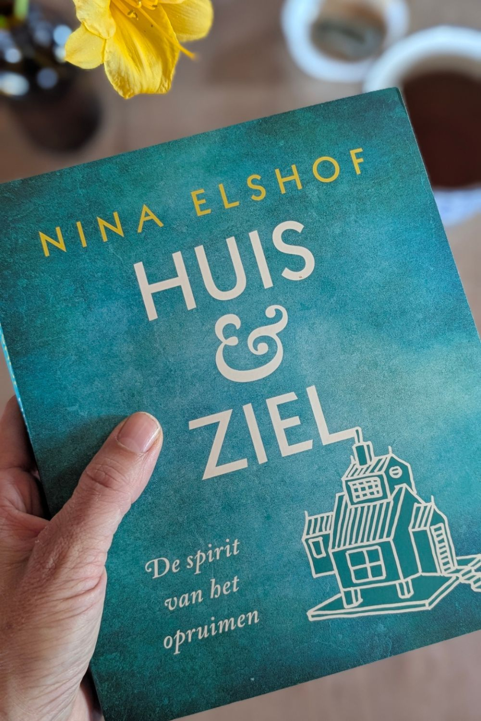 Hand houdt een boek vast met de titel 'Huis & Ziel' en in gele letters de naam van de auteur Nina Elshof