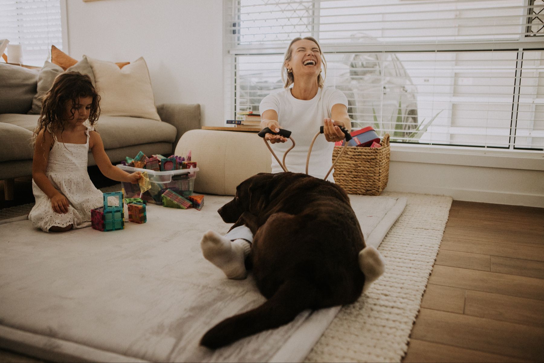 Mum in a lounge room setting laughing with her chocolate Labrador sitting on her as she is using an exercise pilates reformer band her daughter playing with blocks beside her