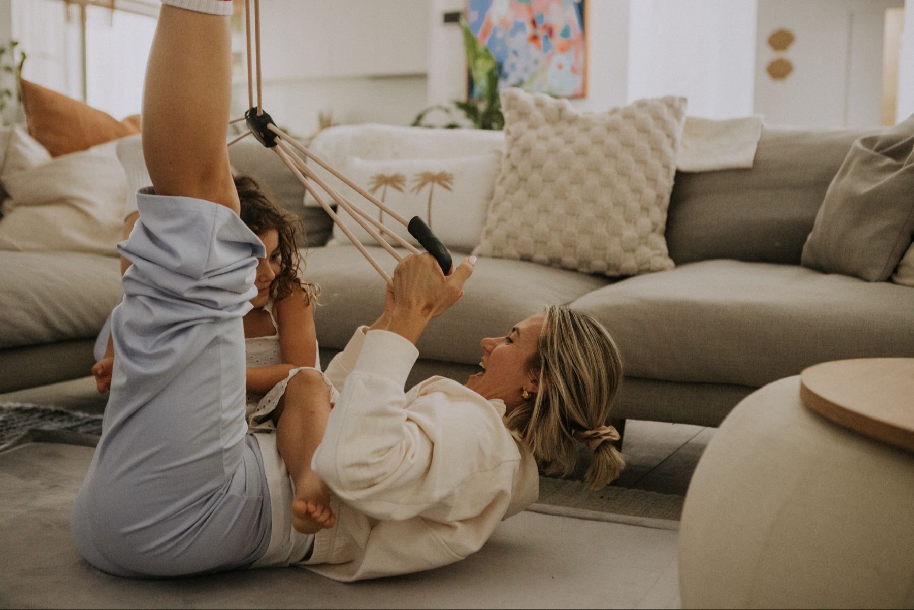 A Mum in casual wear using a pilates reformer exercise band with her legs up and her child climbing on top of her smiling