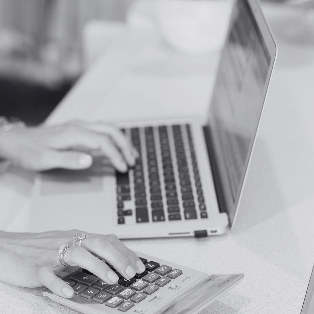 A close-up of a person's hands using a laptop and a calculator simultaneously on a light-colored desk, highlighting a focused workspace for financial planning or business management.