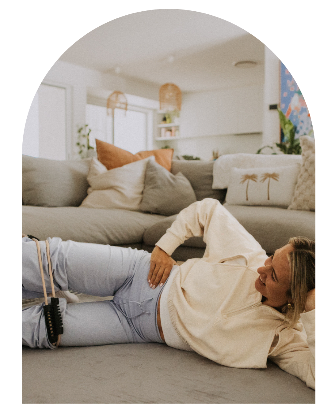 A woman in casual wear in a lounge room working out using a pilates reformer band wrapped around her legs