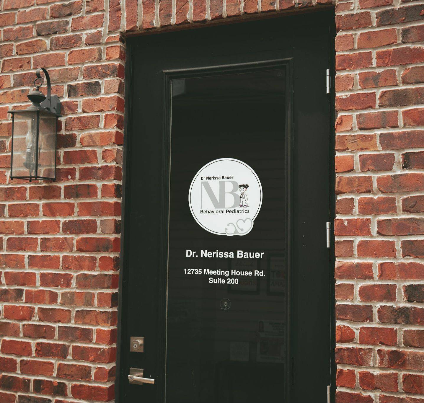 An image of the NB Behavioral Pediatrics front door. It is a black door with the NB Behavioral Pediatrics logo on it. The door is surrounded by red brick and there's a light on the left-hand side.