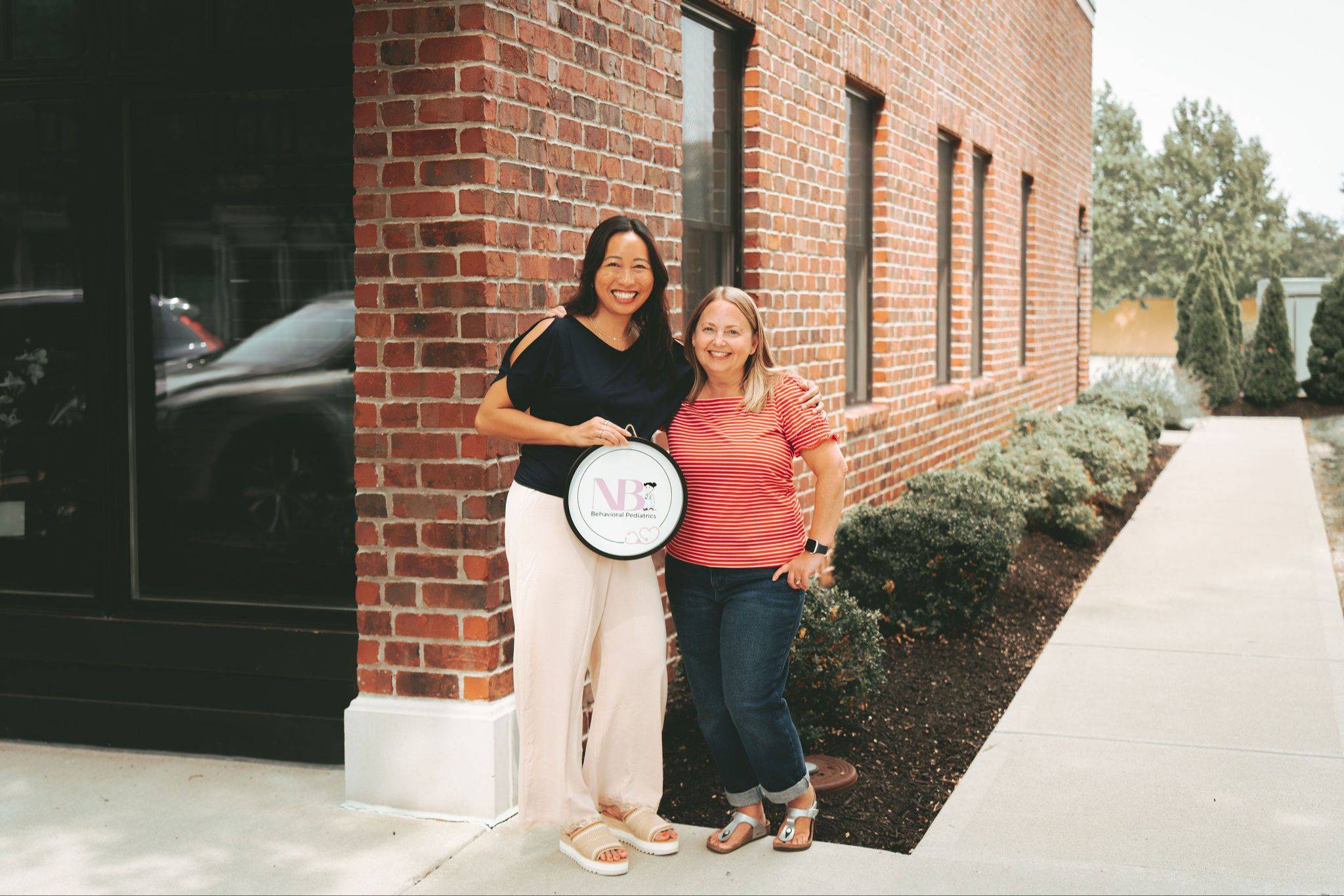 Dr. Bauer and Amy stand in front of the NB Behavioral Pediatrics building. Dr Bauer is holding a sign that has the NB Behavioral Pediatrics logo on it.