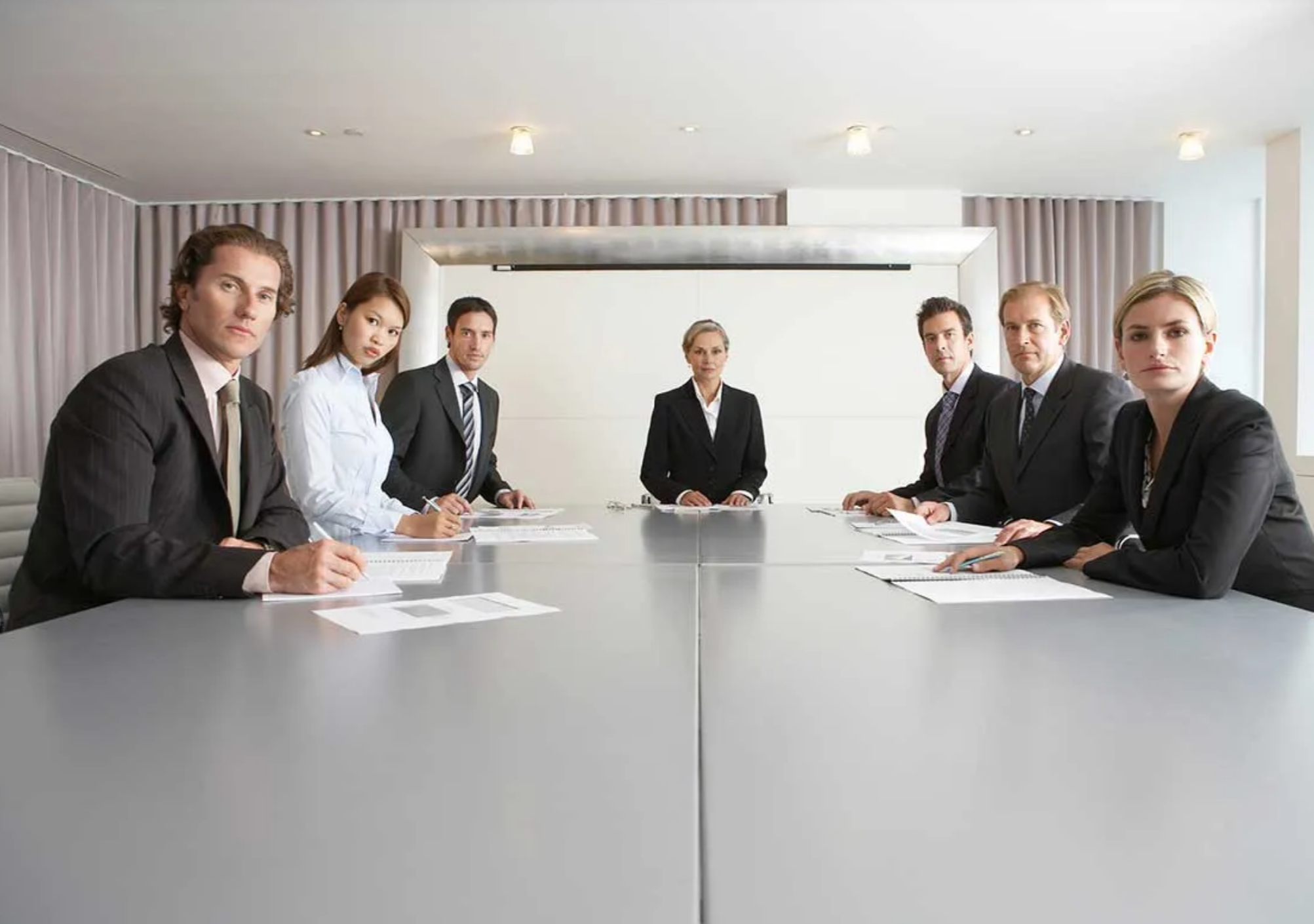 Seven senior executives seated around a boardroom table, looking directly at the viewer