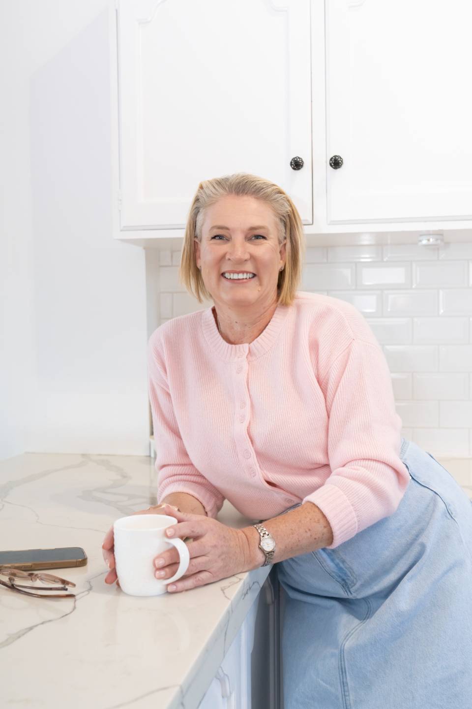 Erin Davis smiling at a kitchen bench with a cup, wearing a pink cardigan at the Dudley shoot