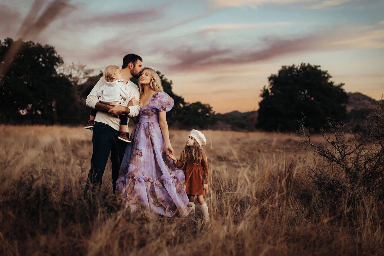 A mother in a lilac dress and a father holding their little ones in a field at sunset