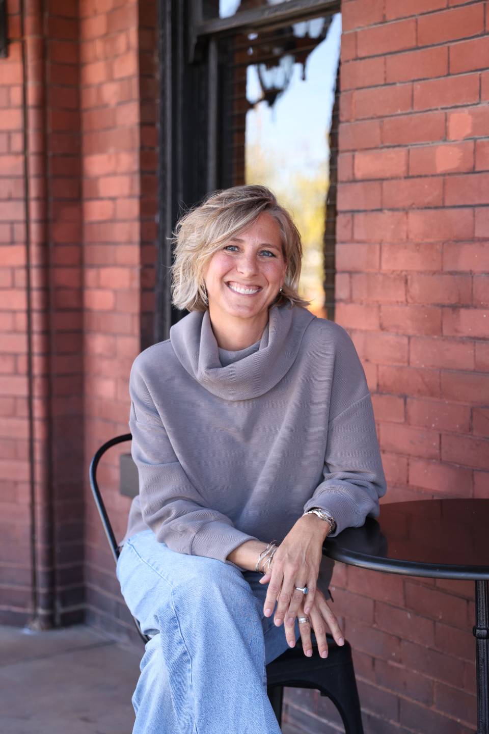 Ciara Stockeland smiling, seated at a bistro table in front of a red brick wall.
