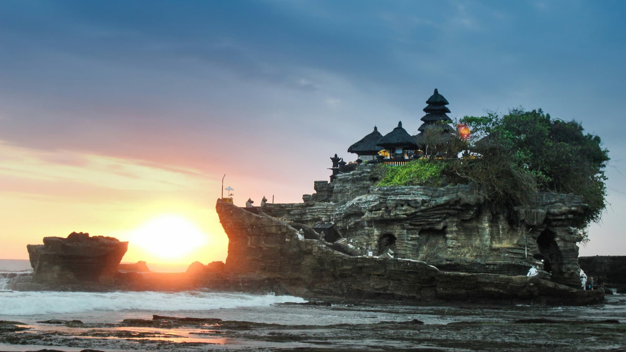 Tanah Lot Temple in Bali at sunset with waves crashing against the rocky shoreline.