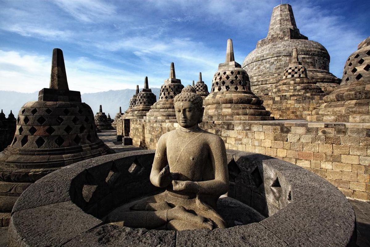 Buddha statue and stupas at Borobudur Temple in Central Java under a clear blue sky.