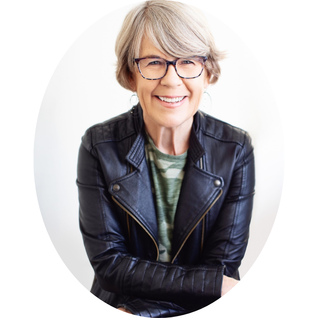 Suzette Mullen, white woman with short blonde hair and glasses sitting cross-legged and smiling