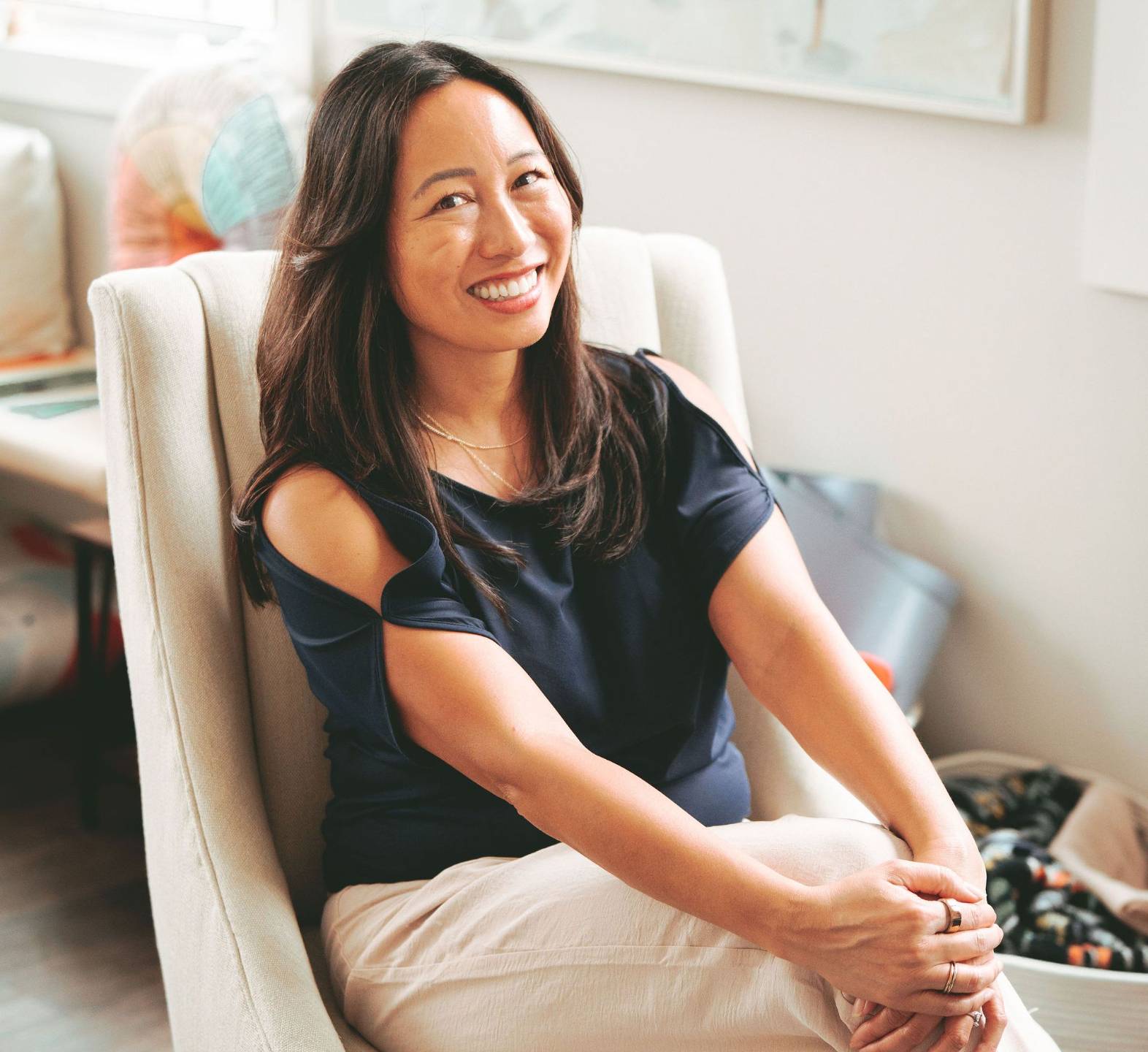 Dr. Bauer is sitting on a cream chair in her office with her hands resting on her right knee.