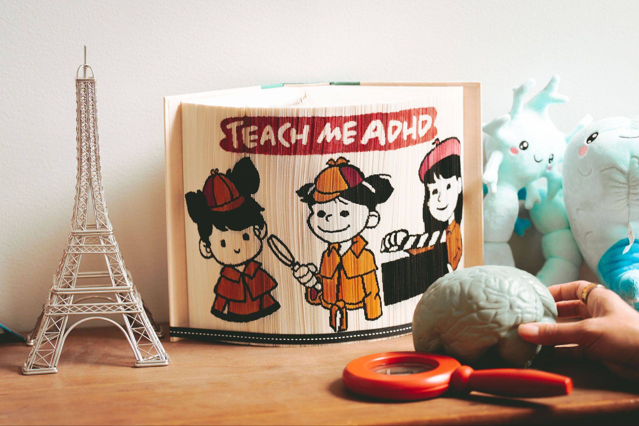 A miniature Eiffel Tower, a magnifying glass, a model brain and an open book with TEACH ME ADHD written on the edges of the pages are all sitting on a wooden surface. A hand is setting the model brain down.