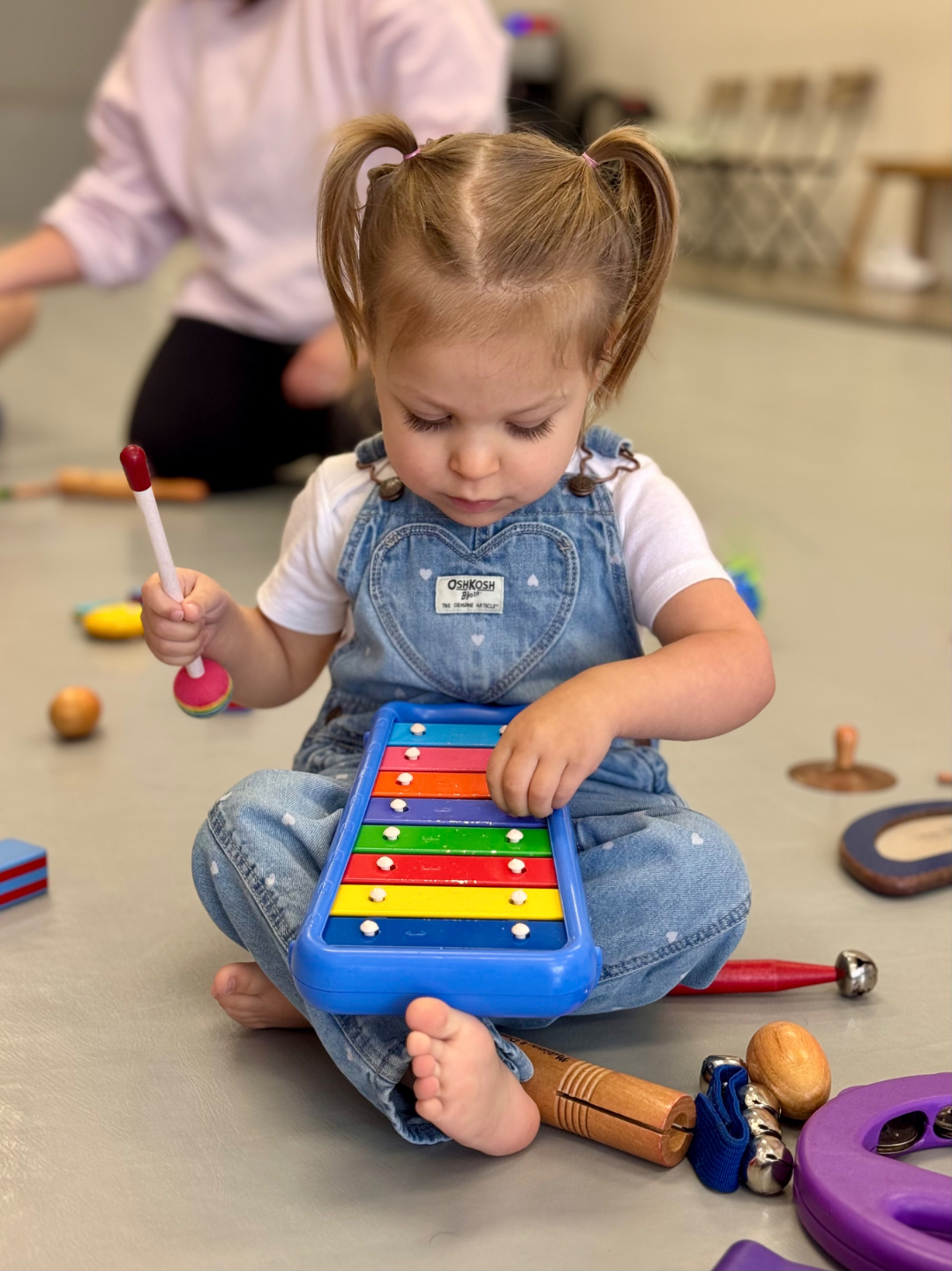young toddler boy plays with simple rhythm instruments while adults sit in background