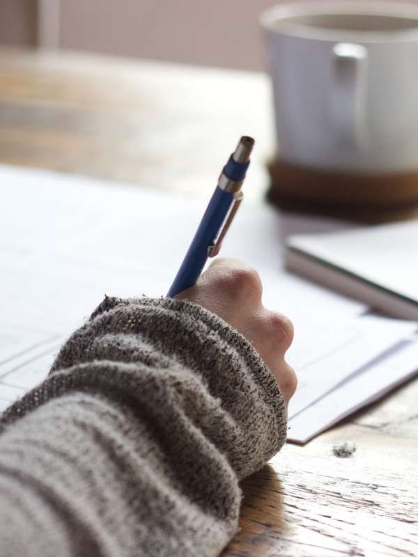 An outstretched hand in a grey jumper holds a blue pen writing on paper