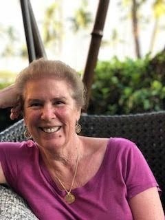 Linda Evans Delman smiles and sits with right hand against her hand in front of natural background