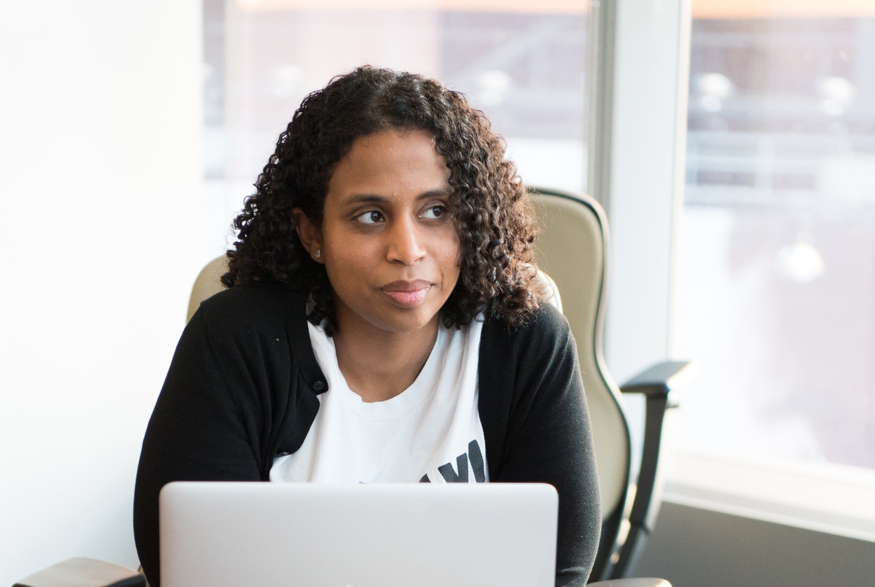 Black woman sitting in front of a computer looking towards the windows. Original Photo by Christina @ wocintechchat.com on Unsplash 