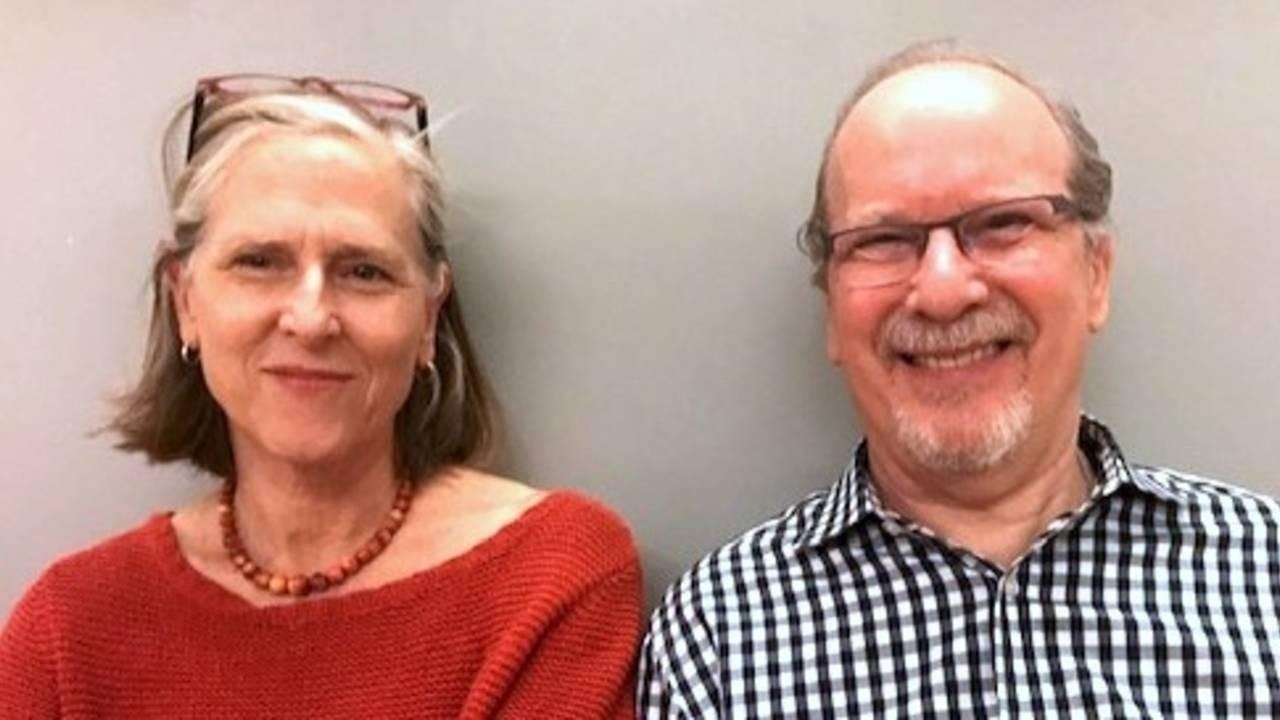 Elizabeth Beringer and David Zemach-Bersin sitting shoulder to shoulder smiling in front of a gray background