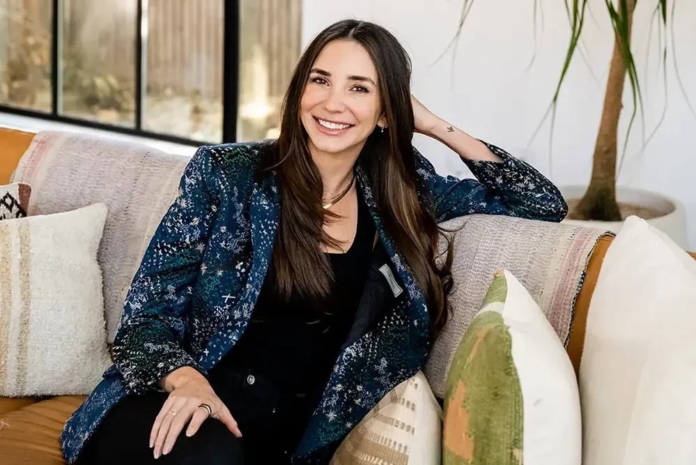 Blaine in a blue blazer, seated on a white couch, smiling and looking into the camera