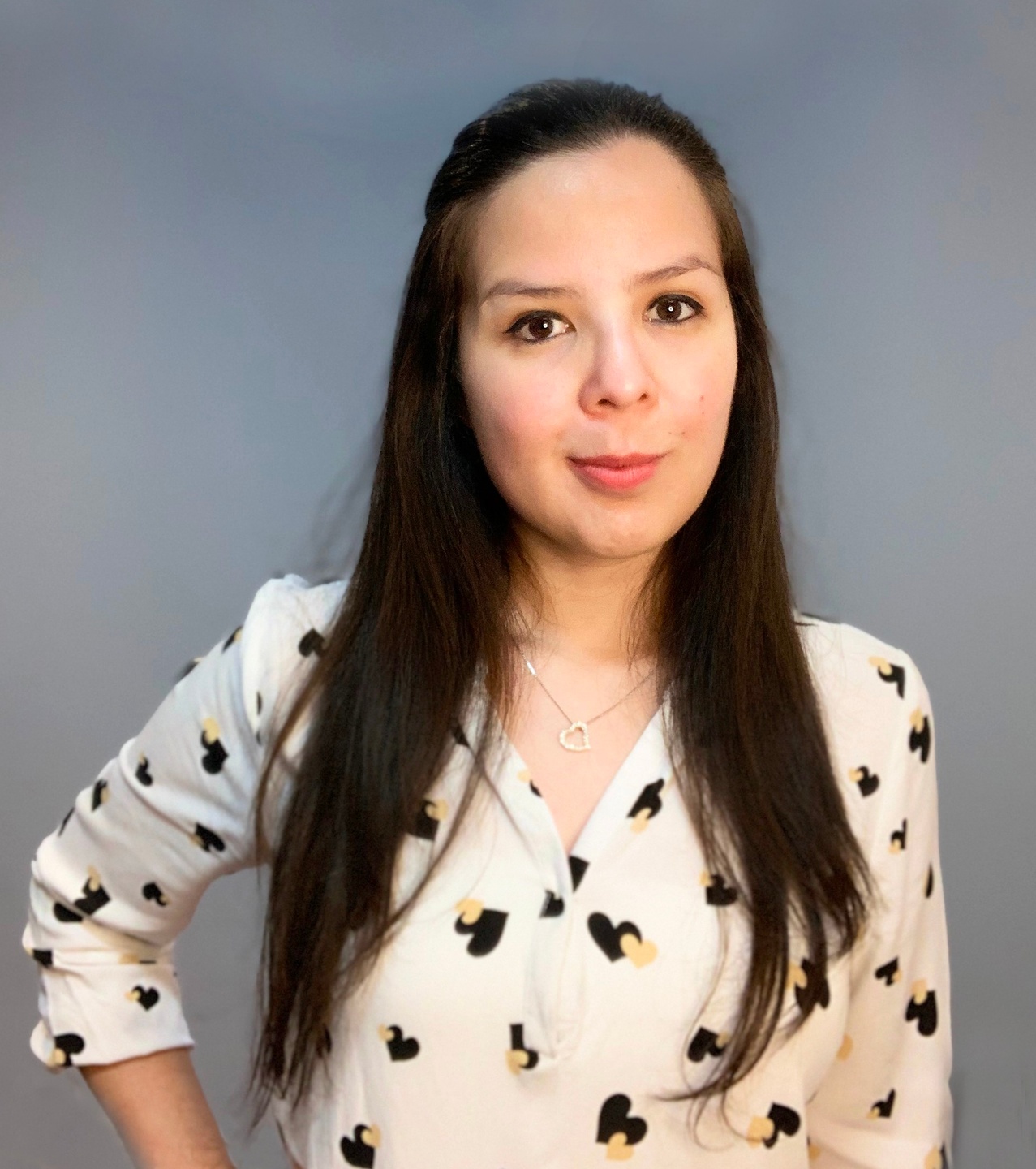 Profile image of Ana Ramirez, a light skinned Latinx woman with long dark hair. She is looking at the viewer and smiling while wearing a white shirt patterned with black and yellow hearts, and a necklace with a heart shaped pendant.