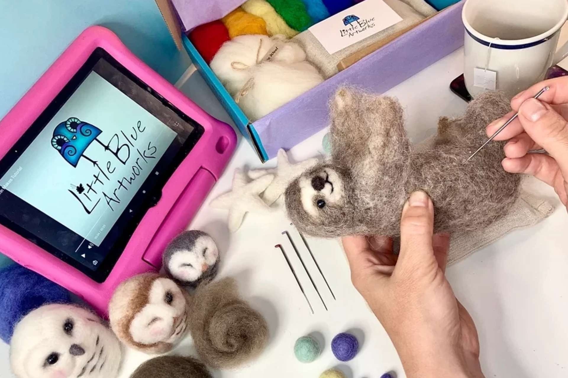 Two hands work on a needle felt sloth, while watching a tutorial video and with all the contents of the kit