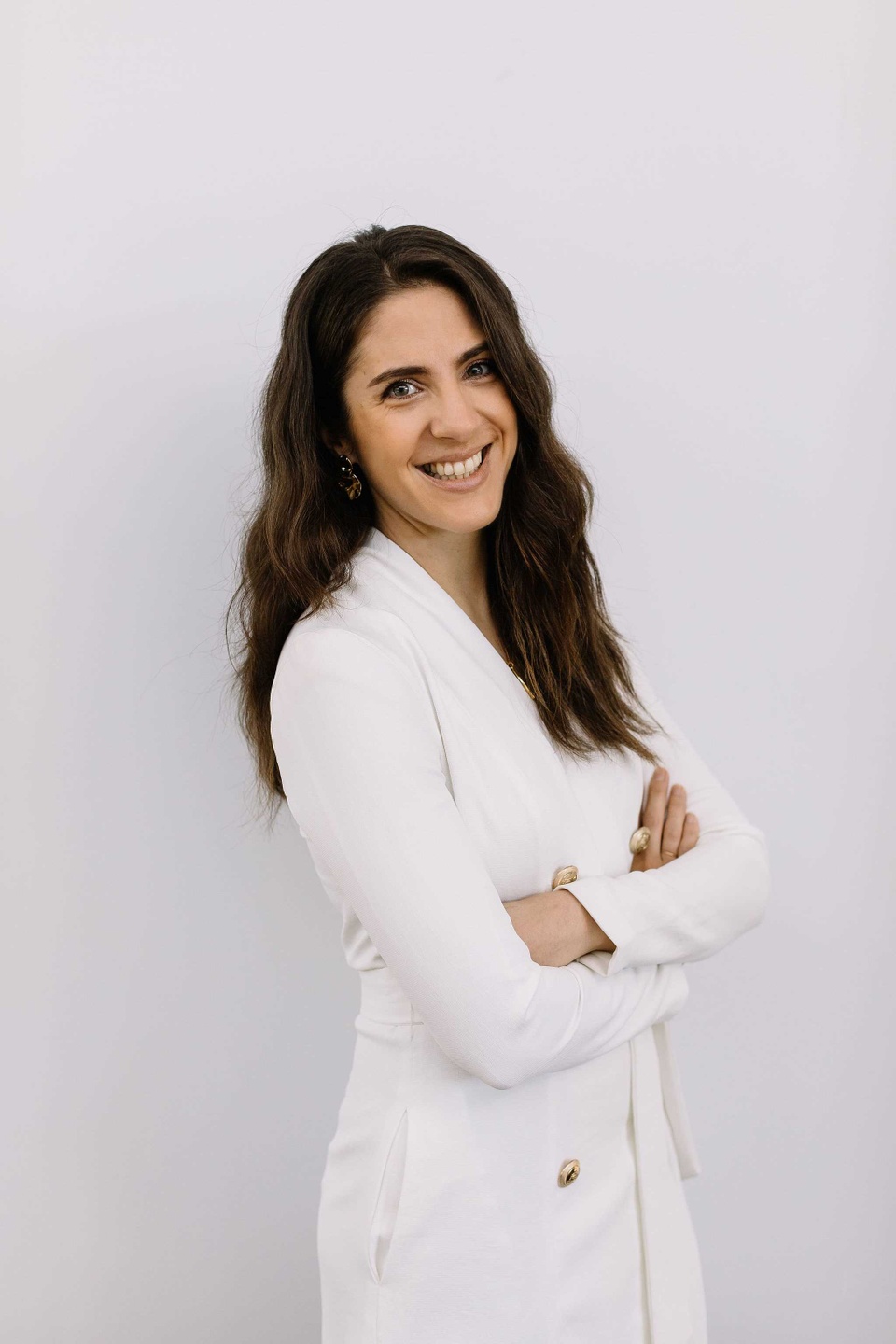 Hareta standing with arms crossed wearing white and smiling at camera in front of white wall