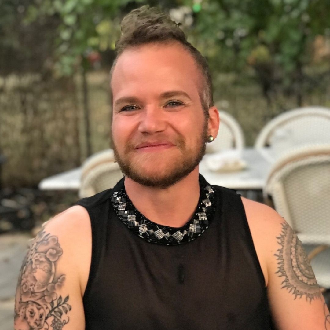 A photo of Kelly, a white transmasculine non-binary person sitting in a chair with plants behind them. They are smiling into the camera and wearing a black tank. They have tattoos on their arms and they are wearing eye makeup. 