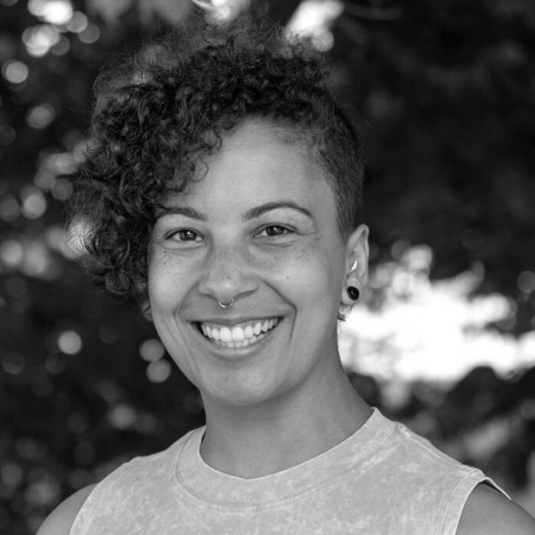 A photo of Kelly, a white transmasculine non-binary person sitting in a chair with plants behind them. They are smiling into the camera and wearing a black tank. They have tattoos on their arms and they are wearing eye makeup. 