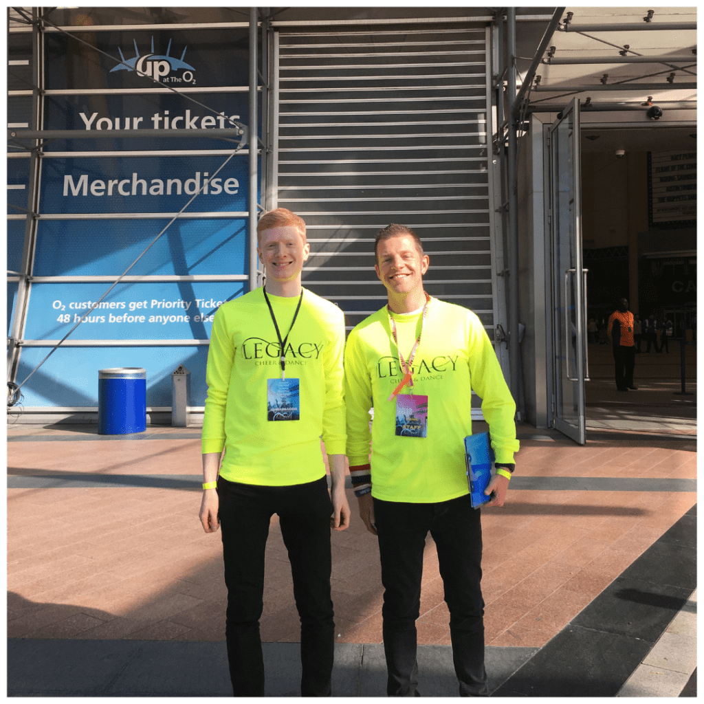 Two young men in bright neon Legacy shirts smile warmly, welcoming guests to the venue